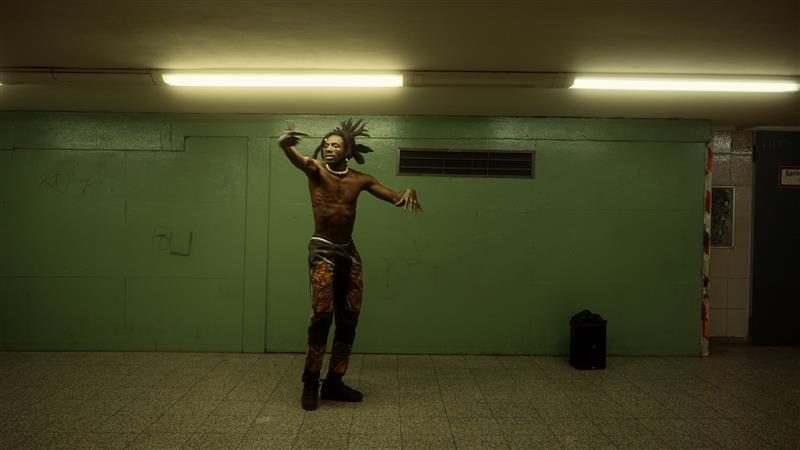 A shirtless man dances in a subway station infront of a green wall and below fluorescent lighting.