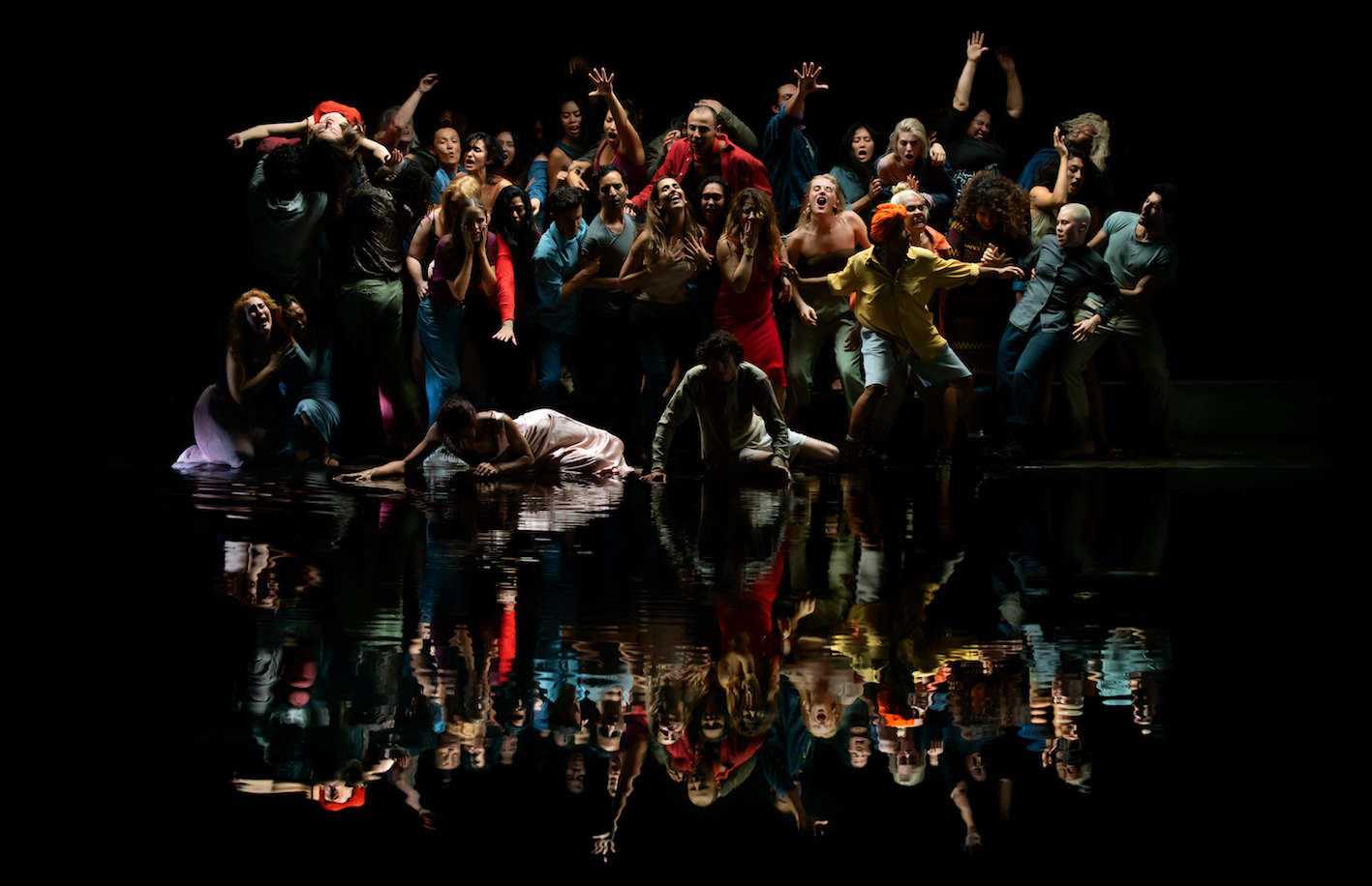 A large group of people in colourful clothing pose infront of a body of water with a black backdrop.