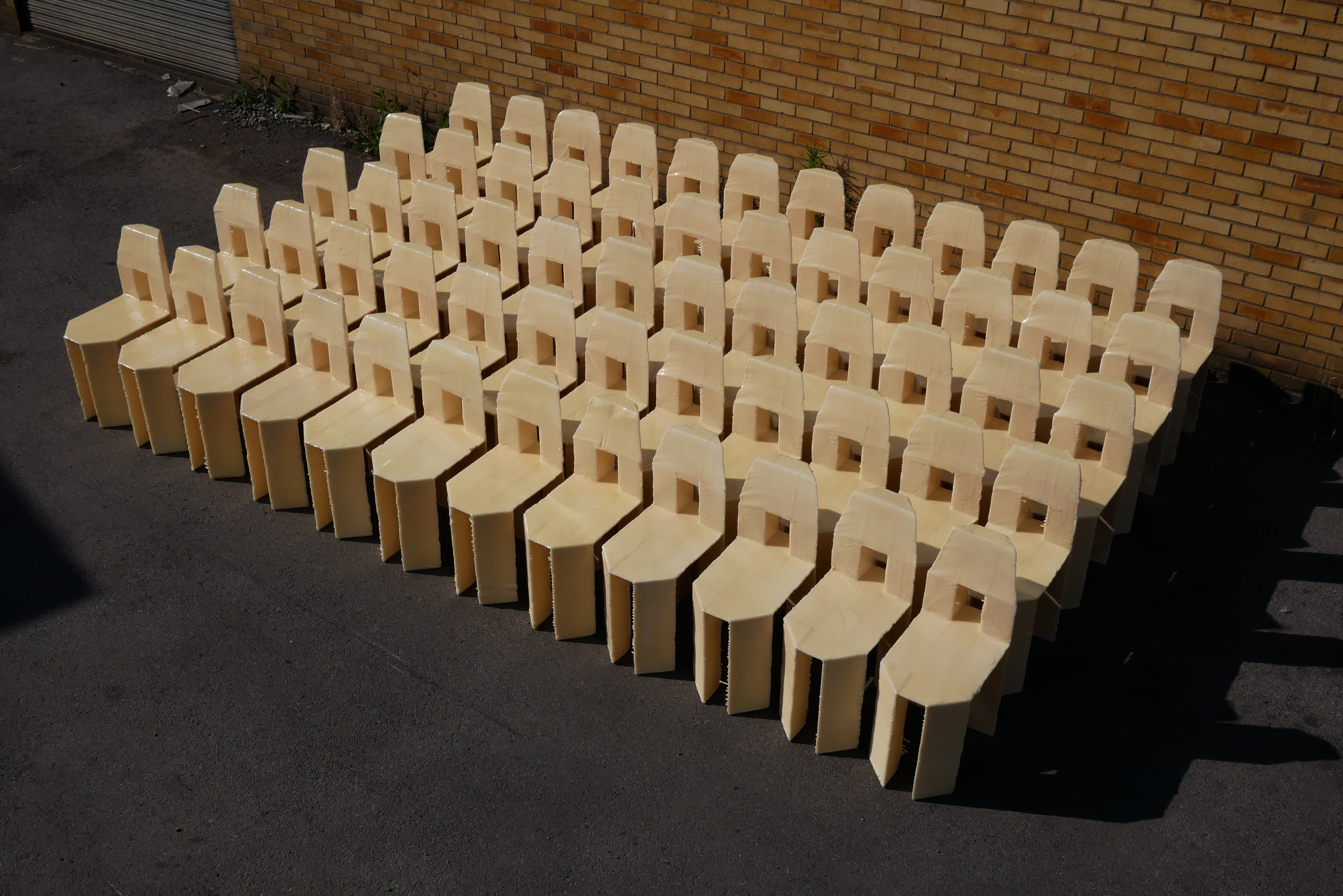 A group of 60 cream chairs with a square cut out in the back sit angled to the left in the sun infront of a brown bricked wall.