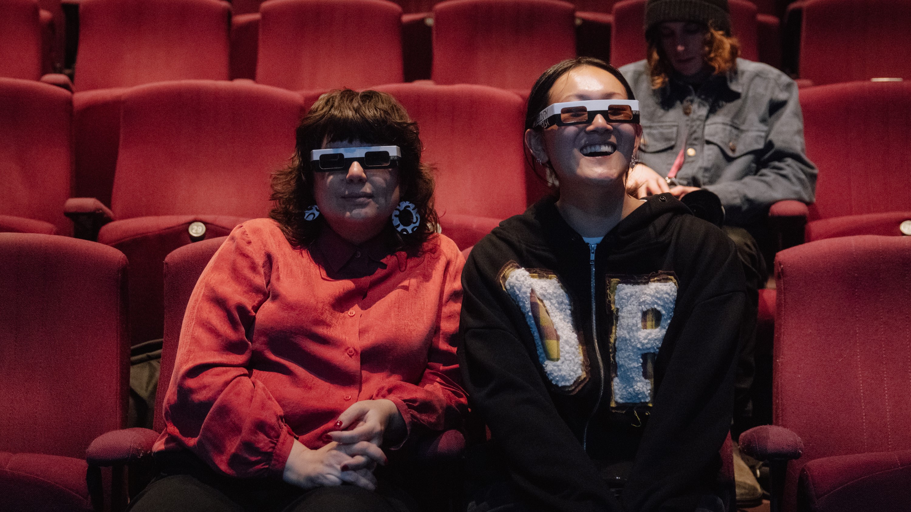 Two women wearing 3D glasses sit in an auditorium, intended to block out the AI-generated dance cues.