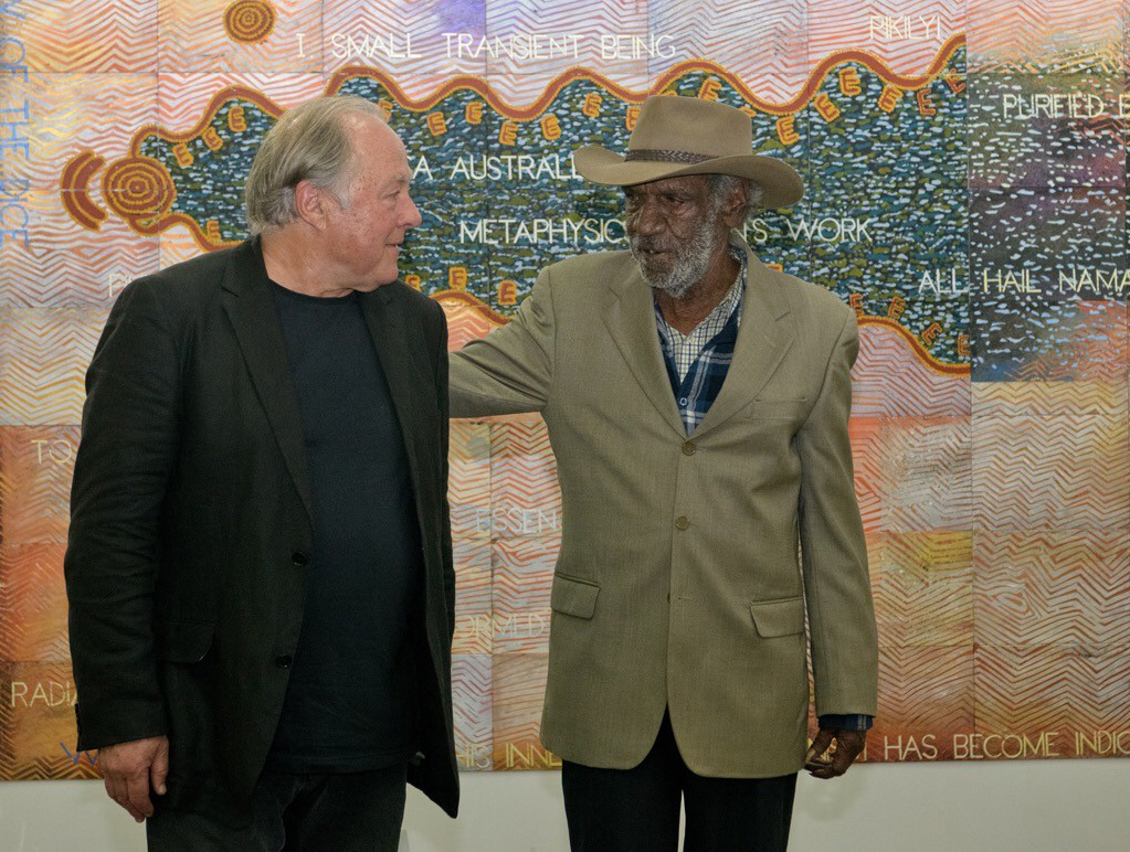 Two men looking at each other, one with his arm on the other's back, standing in front of a pastel coloured artwork.