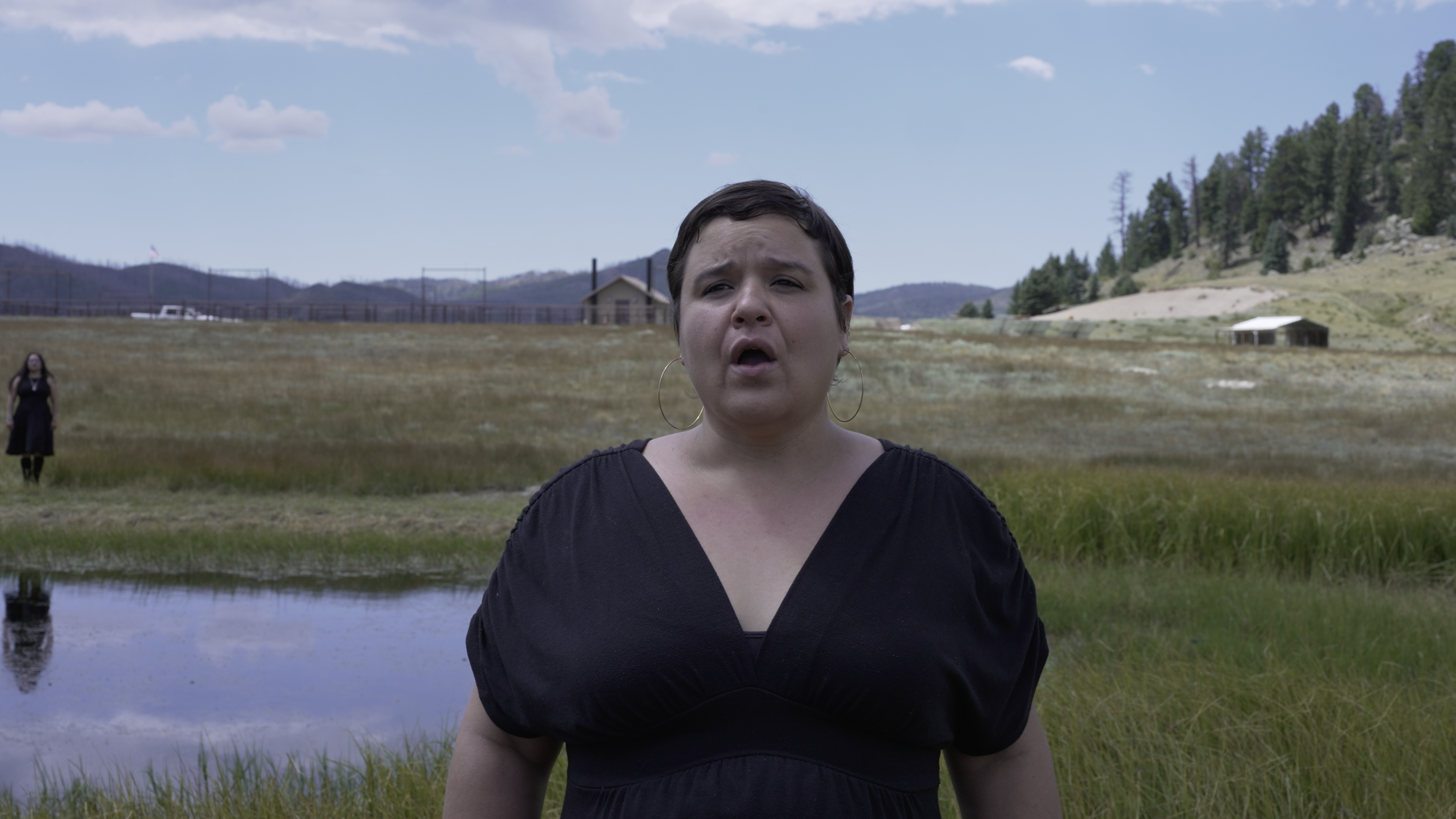 A white woman with short dark hair wearin a black dress stands in a field with a body of water behind her.