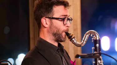 A man wearing glasses and a dark suit plays a saxophone.