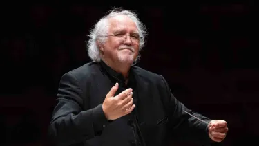 An older man with white hair, wearing glasses and a black suit, stands conducting with a baton in his right hand.