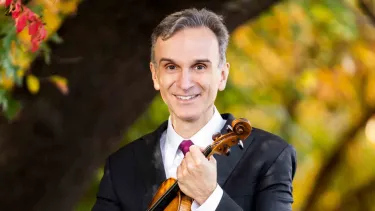 Gil Shaham holds a violin in a green forest.