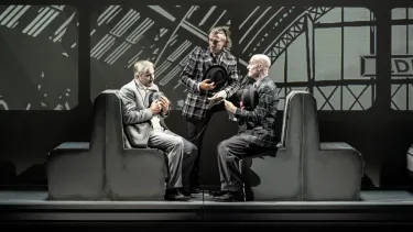 A group of three people sitting in facing each other on upright wooden bench seating. All holding felt hats.