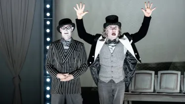 Two people stand side by side, one wearing a top hat, with both hands raised above their head.