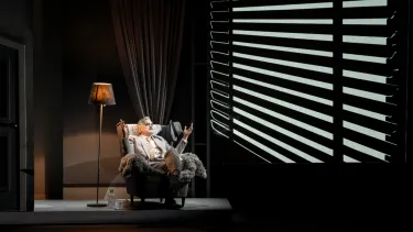A people sitting in a darkened room on an armchair, with both arms raised as if in contemplation.