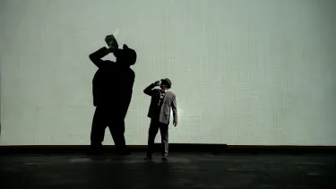 A person standing on an empty stage drinking from a glass, with their shadow cast in the background.