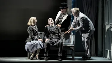 Four people on stage, two sitting on a wooden bench, two standing behind and to the side. All in mid-conversation.