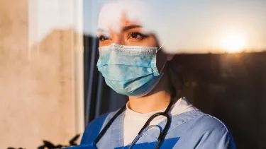 A doctor wearing a mask and a statoscope looking through a window.