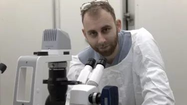 Dr Brett Kagan in a white labcoat standing over a microscope looks at camera