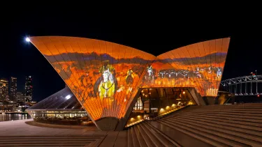 The Sydney Opera House sails at night, illuminated with an Australian outback landscape in orange and browns with a blue sky.
