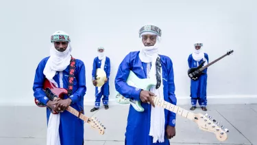 Four men wearing blue outfits and white headscarves, three holding a guitar and one holding a drum cymbal.