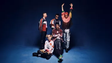 A group of five men dressed in kooky, punky Y2K outfits pose in front of a blue backdrop.