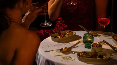 A close up of a round table with a white table cloth filled with canapes and cocktails for the VIP Gatsby at The Green Light experience, with moody, candle lit lighting.