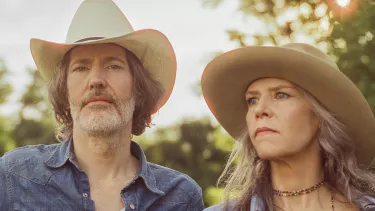 A man holding a guitar and woman both in cowboy hats look pensive away from the camera exuding a country music vibe.