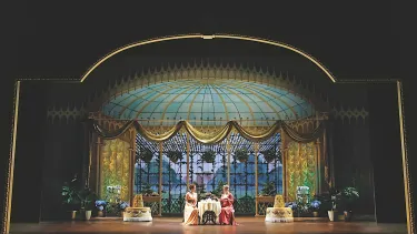 Eliza drinks tea in a palacial glass room.