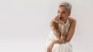 A woman adorned with tattoos with short, bleached blonde hair is comfortably positioned on a chair, resting her chin in her hand.