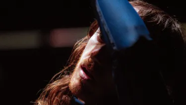 A man with long hair poses thoughtfully mid-movement, he wipes his forehead with his forearm.