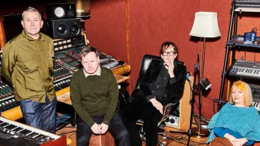 Three men and one woman in their 60s sit in a music recording studio.
