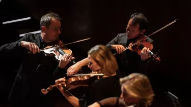 A 50-year old man plays violin next to another male violinist and a female violinist.