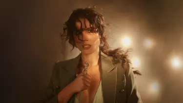 A white woman with a messy, curly ponytail and wearing a beige blazer looks worried at the camera while holding the handle of a sword.