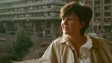 A woman resting her arm on a wall wearing a beige long coat.