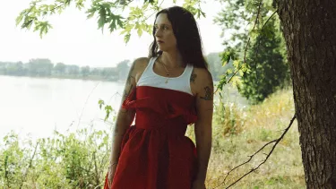 Woman in red dress standing by river, gazing into distance.