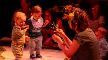 A women wearing a crown of flowers is holding up some leaves to two toddlers.