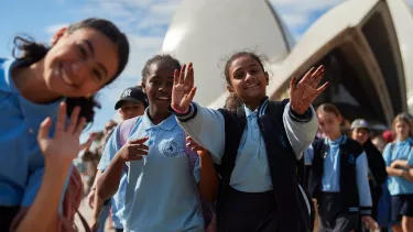 A group of girls wave at the camera in school uniform. The Sydney Opera House sails are seen blurry in the background.