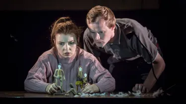 A man and a woman stare intently at small puppets of people.