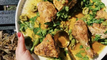 Roasted sheet pan chicken with crushed olives presented in a white bowl by a person standing outside. A brass spoon is resting on the plate.