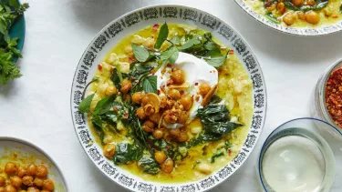 A white ceramic bowl with black ornate edge pattern holding spiced chickpea stew with coconut and turmeric on a tabletop. Five smaller bowls surround the edge of frame holding ingredients.