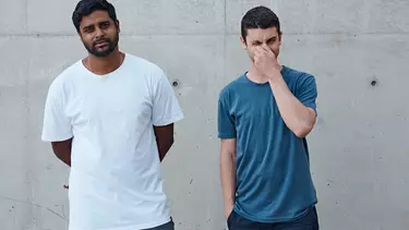 A man in a white T-shirt next to a man in a blue T-shirt smiling against a wall.