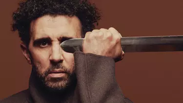 A man with stubble beard and curly hairs holding a sword blade.