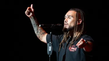 A man with dreadlocks delivering a slam poem with his arm in the air.