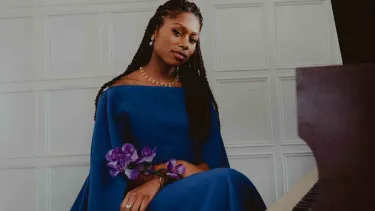 Isata Kanneh-Mason pictured in front of a grand piano in a blue dress.
