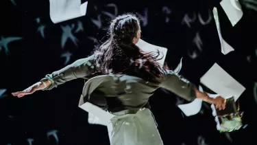 A women dances surrounded by falling paper. She is holding a book and her hair is in braids.