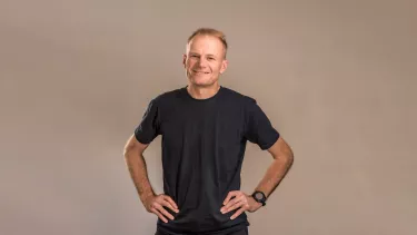 A man with short light brown hair smiles with his hands on his hips. He wears a black t-shirt.