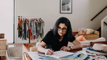 A woman seated in a house, drawing into a notebook at a desk.