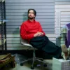 A white man sits in a chair with his feet up wearing a red sweater.