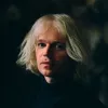 A white man in early 40s with jaw-length white hair looks at the camera.