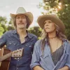 A man holding a guitar and woman both in cowboy hats look pensive away from the camera exuding a country music vibe.