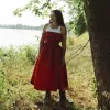 Woman in red dress standing by river, gazing into distance.