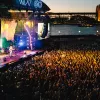 A large crowd stands in front of an outdoor stage at sunset with the Sydney Harbour Bridge in the background.