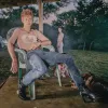 A white man wearing a beige top and baggy jeans louges in a plastic chair.