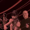 A Singaporean man and woman play the cello and violin respectively next to an older white man holding a conductor's baton, all basked in a red light filter.