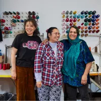 A group of three women standing together in a room with colourful spools hanging on the wall behind them.