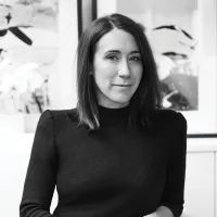 Black and white portrait of a woman with shoulder-length hair in a dark top, standing in front of abstract art. Her expression is calm and thoughtful.